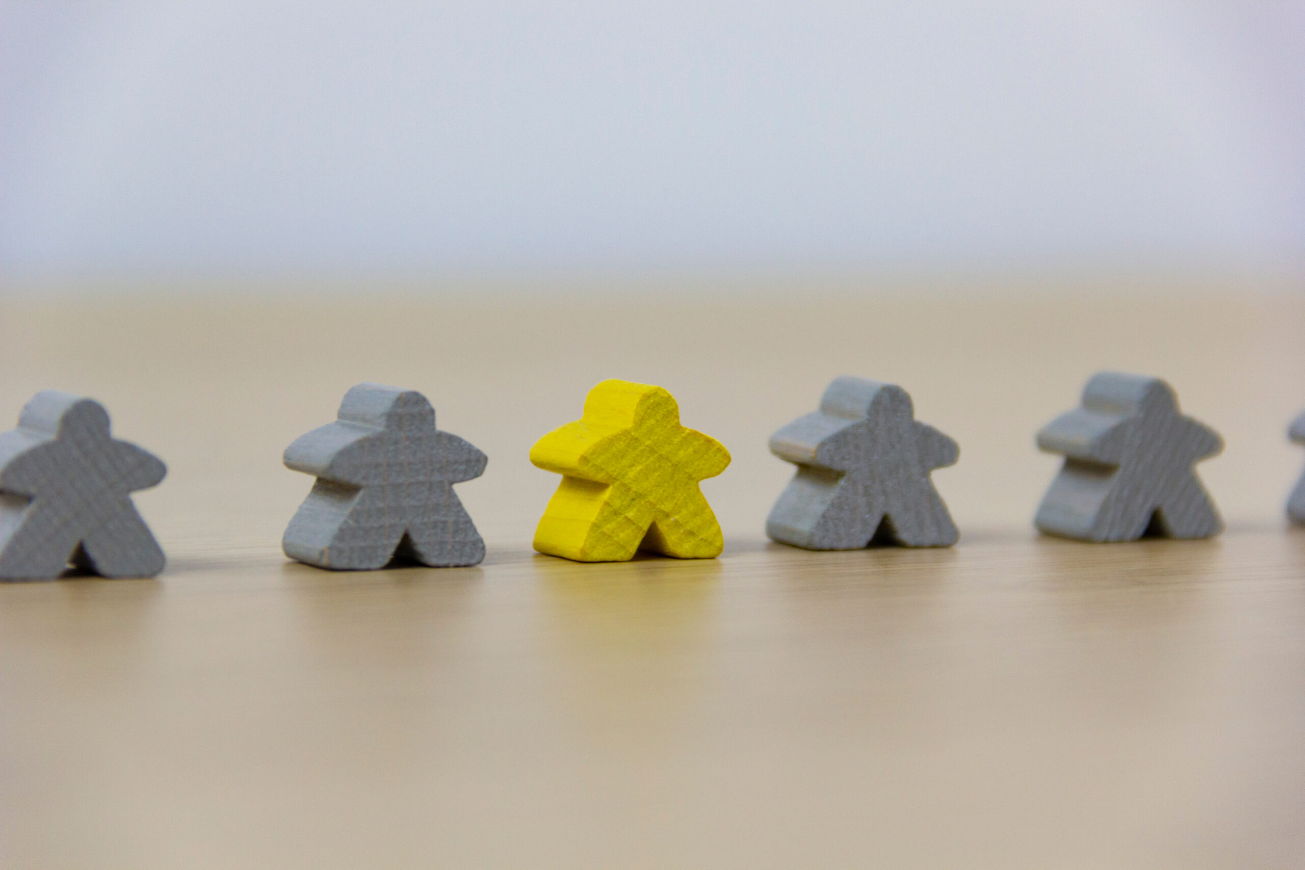 Several grey wooden meeple figures standing in a row on a light wooden surface with one distinct, bright yellow meeple figure in the center, symbolizing leadership, individuality, or standing out in a team.